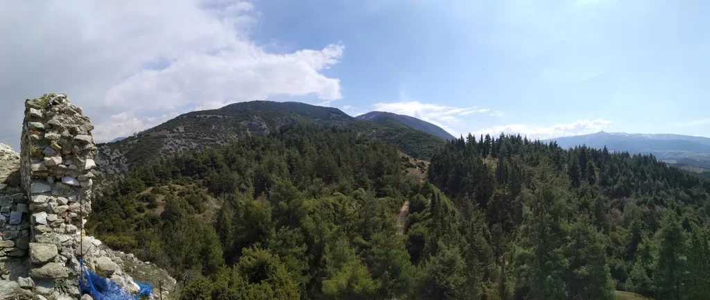 Φωτογραφία όπου απεικονίζεται το φυσικό ορεινό τοπίο, όπως αυτό διακρίνεται από την ακρόπολη των Σερβίων. Στα αριστερά της φωτογραφίας διακρίνονται ερείπια ενός πύργου της ακρόπολης των Σερβίων και στο κέντρο διακρίνονται βουνά που καλύπτονται από κωνοφόρα δέντρα.